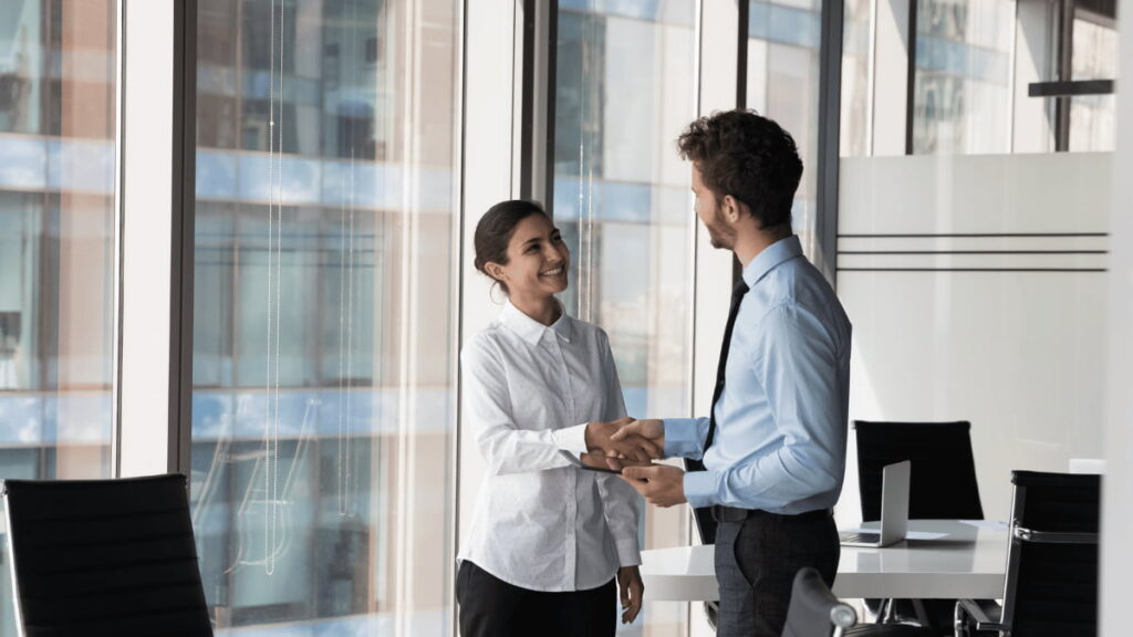 Werkgever en sollicitant schudden elkaar de hand in een modern kantoor met grote ramen en natuurlijk licht.