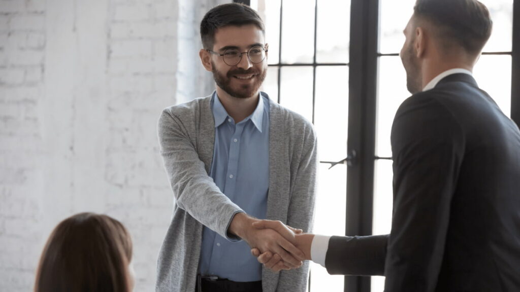 Twee personen schudden elkaar de hand tijdens een sollicitatiegesprek in een modern kantoor met grote ramen en natuurlijk licht.